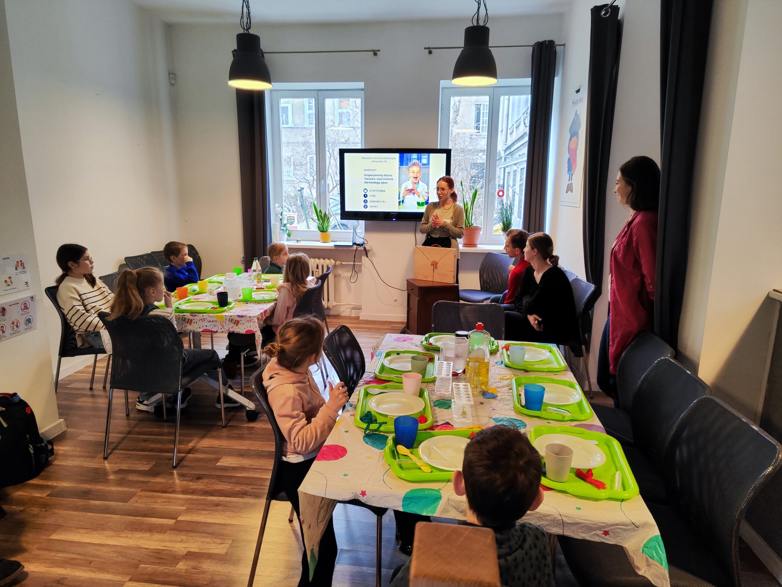 27.01 Warsztaty „Eksperymenty Stasia Taczaka, czyli chemia dla każdego ejbra” W centrum fotografii widzimy panią opowiadającą o zadaniach, a przed nią siedzą dzieci przy stołach.