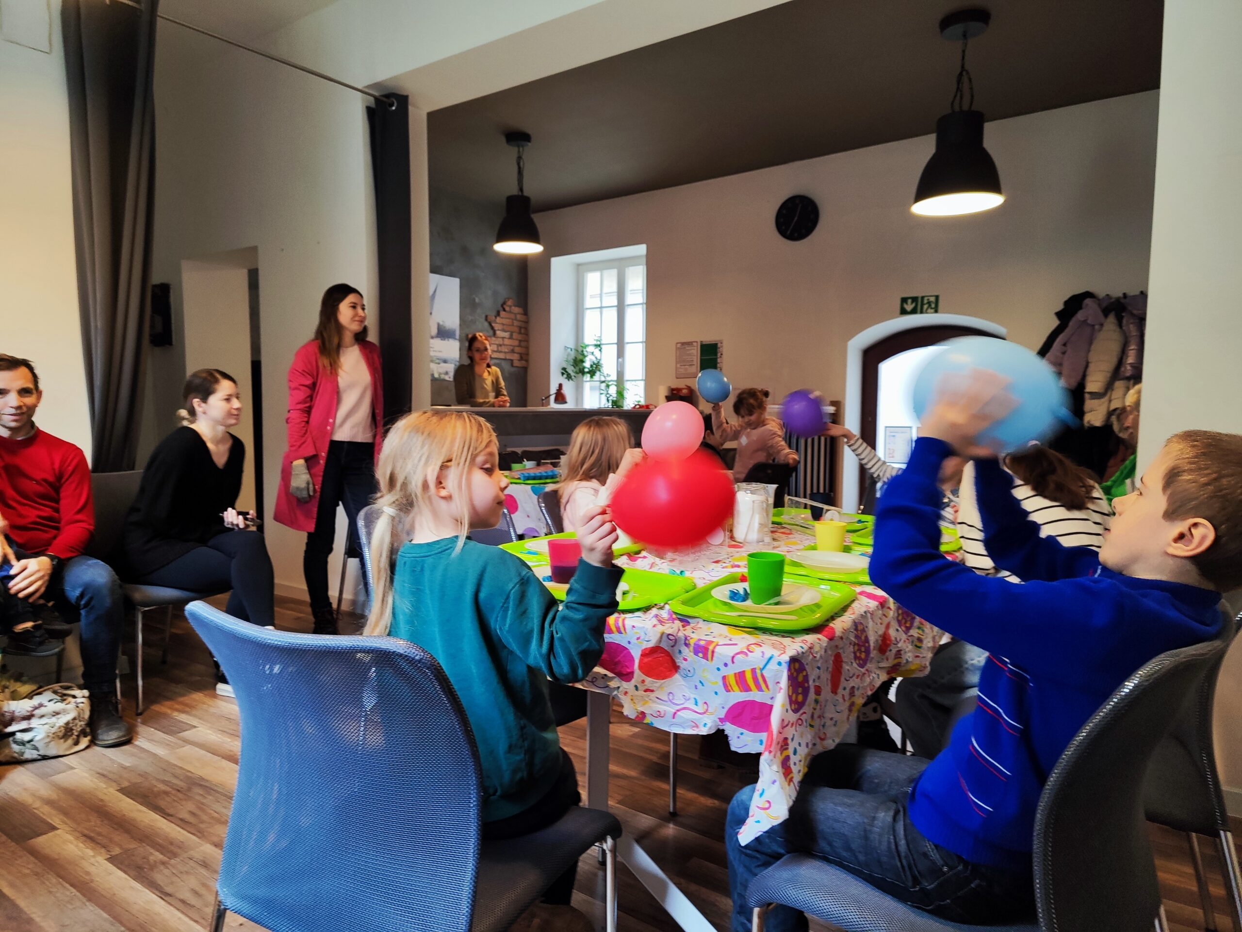 Zdjęcie z warsztatów pod tytułem „Eksperymenty Stasia Taczaka, czyli chemia dla każdego ejbra”. Na fotografii dzieci przy stole bawią się niebieskimi i czerwonymi balonami.