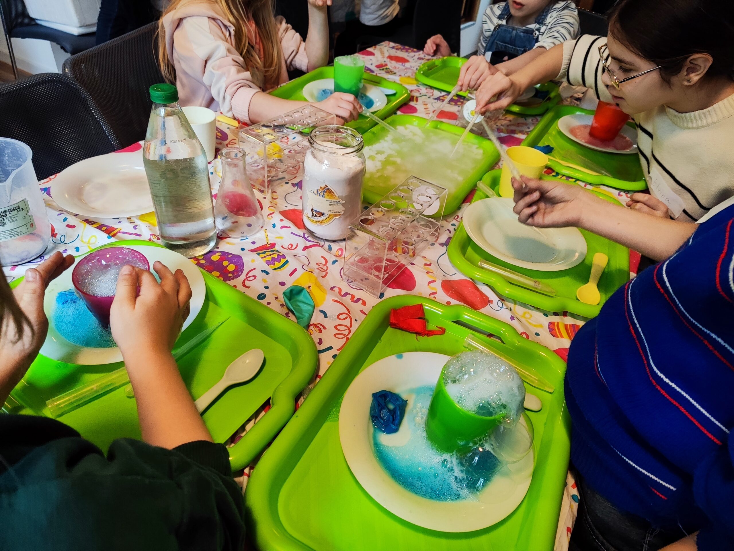 Zdjęcie z warsztatów pod tytułem „Eksperymenty Stasia Taczaka, czyli chemia dla każdego ejbra”. Na fotografii dzieci tworzą pianowe wulkany z płynu do mycia naczyń, sody, octu i suchego lodu.