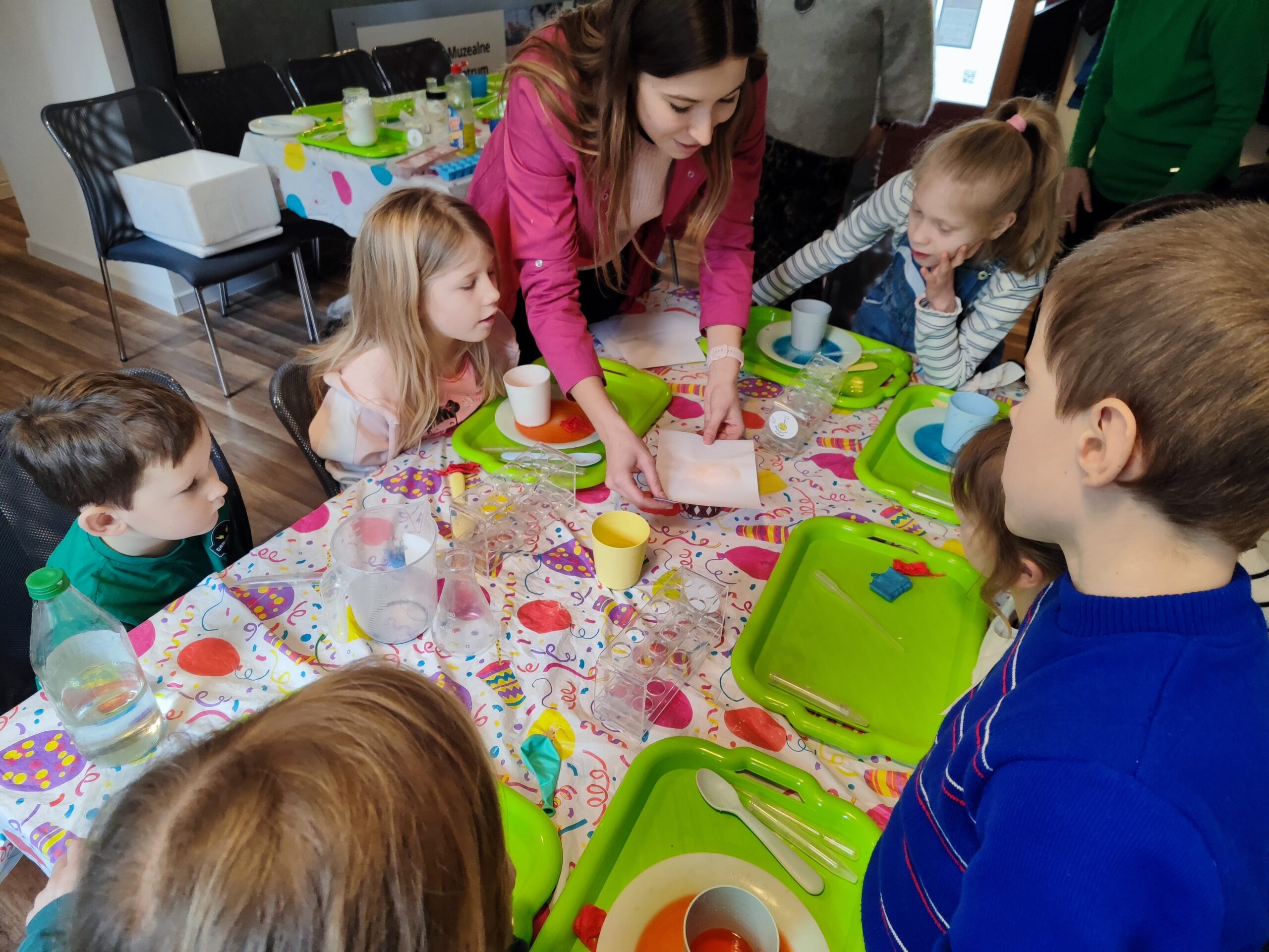 Zdjęcie z warsztatów pod tytułem „Eksperymenty Stasia Taczaka, czyli chemia dla każdego ejbra”. Na fotografii prowadząca pokazuje dzieciom kartkę, a dzieci uważnie słuchają jak tłumaczy.