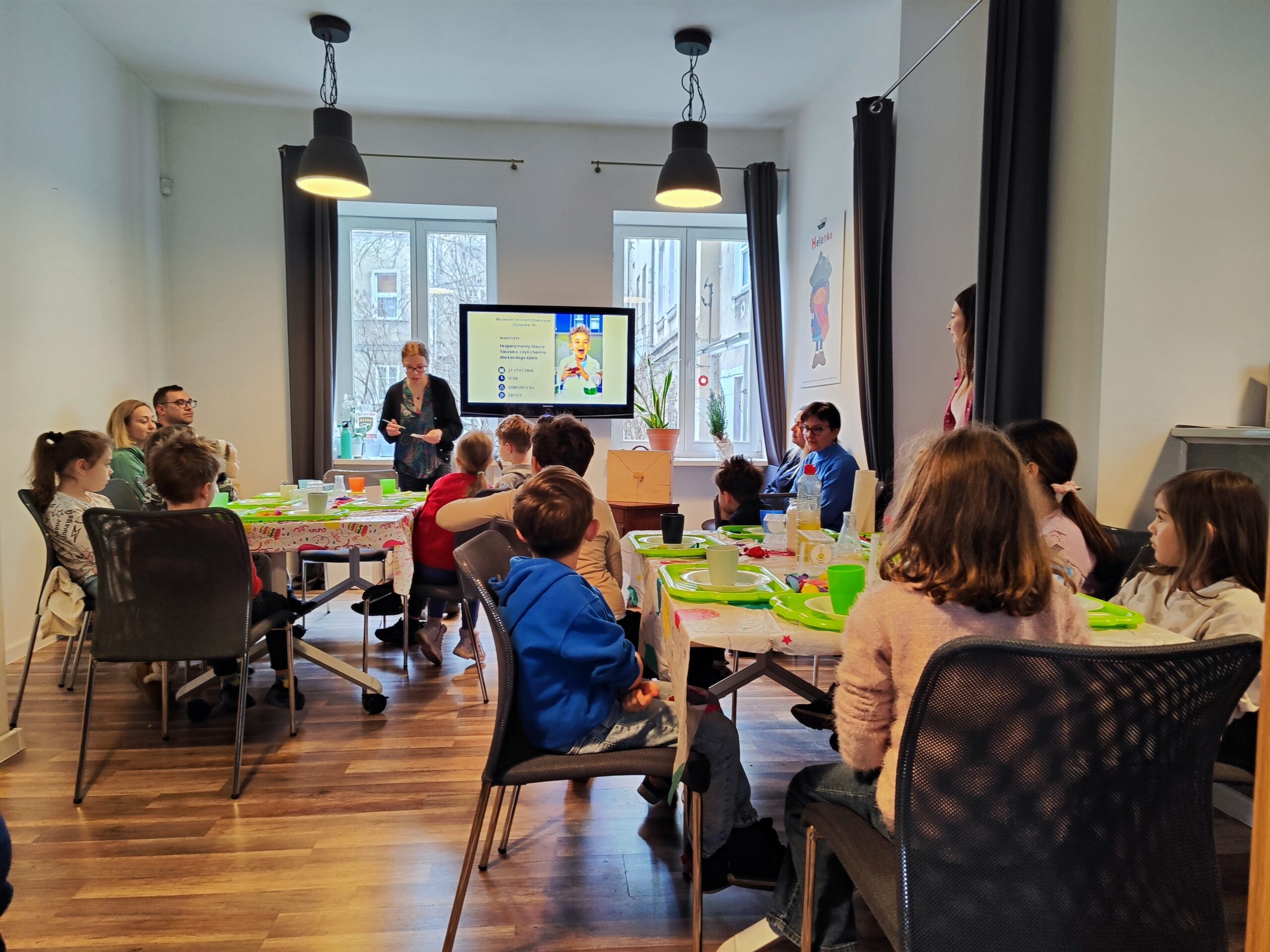 Zdjęcie z warsztatów pod tytułem „Eksperymenty Stasia Taczaka, czyli chemia dla każdego ejbra”. Obraz grup warsztatowych przy stołach, w tle prowadzące zajęcia.
