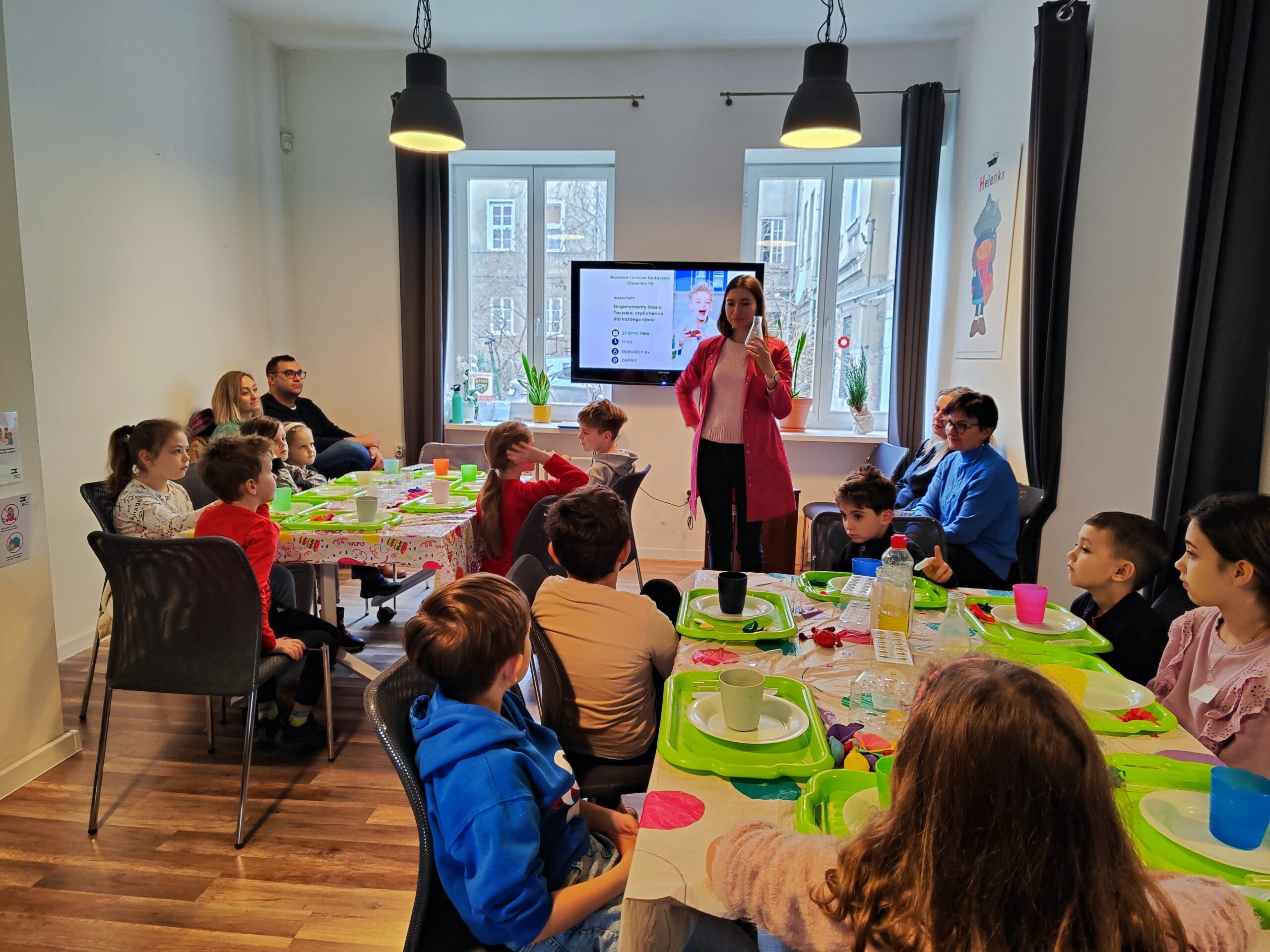 Zdjęcie z warsztatów pod tytułem „Eksperymenty Stasia Taczaka, czyli chemia dla każdego ejbra”. W centrum zdjęcia znajduje się pani tłumacząca dzieciom, co mają zrobić.