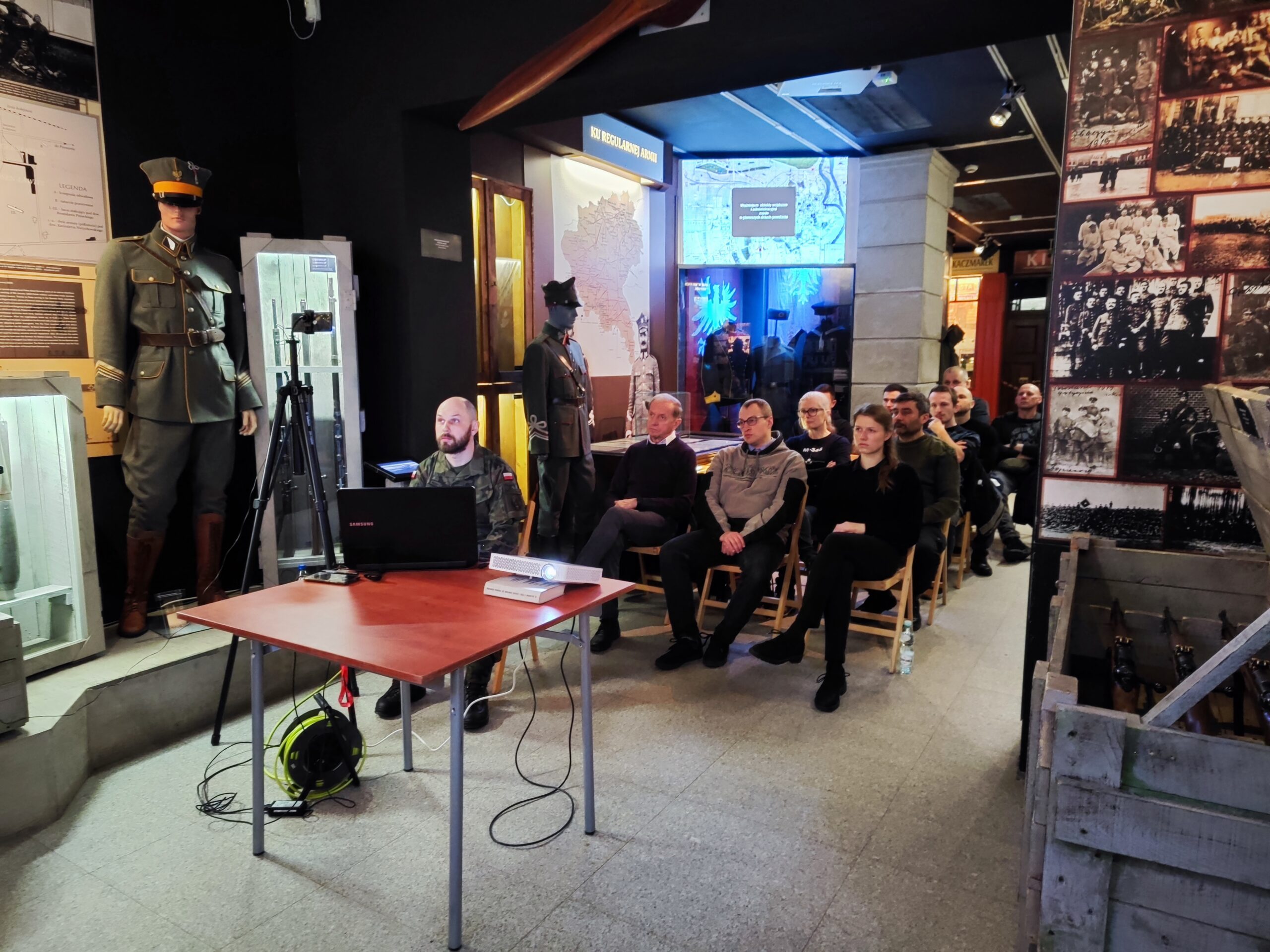 Zdjęcie przedstawia publiczność słuchającą uważnie prelekcji w Muzeum Powstania Wielkopolskiego 1918-1919. Z przodu znajduje się osoba towarzysząca, która zmienia slajdy na prezentacji.