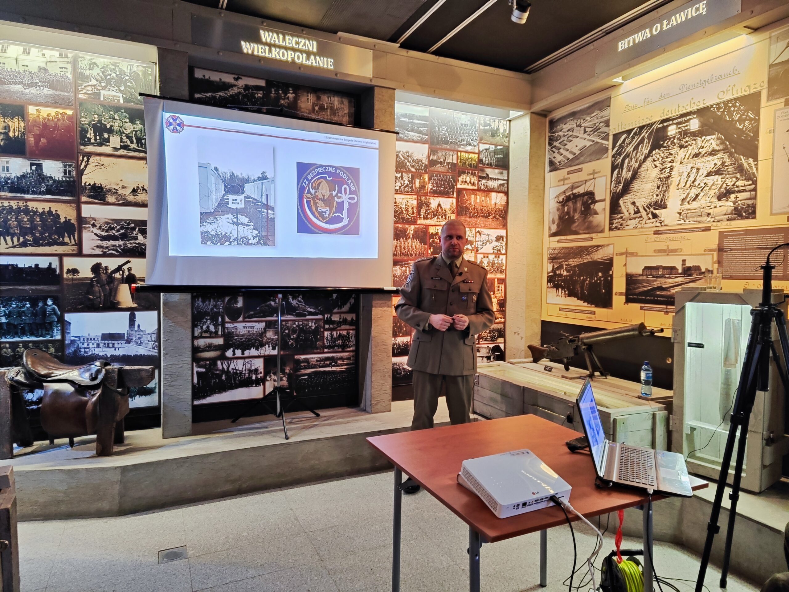 Zdjęcie przedstawia prelekcje w Muzeum Powstania Wielkopolskiego 1918-1919. Na pierwszym planie znajduje się prowadzący z laptopem. W tle wyświetlony jest slajd z prezentacji.