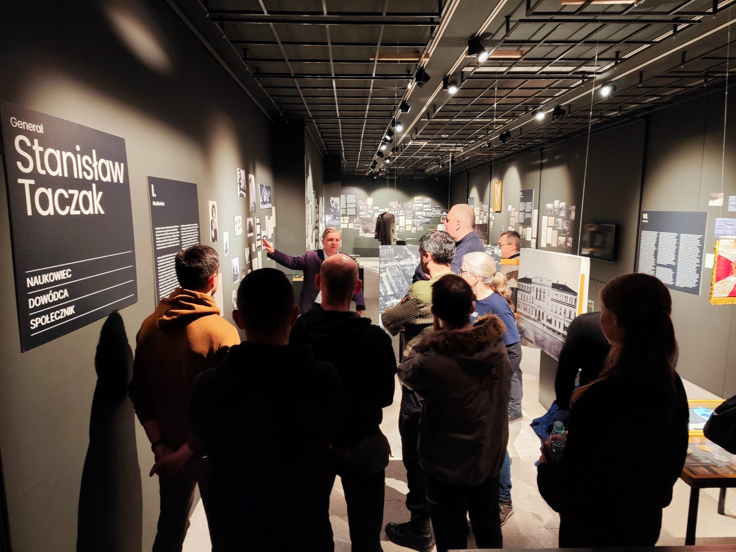 Zdjęcie przedstawia grupę osób zwiedzających wystawę czasową o Stanisławie Taczaku „Naukowiec, dowódca, społecznik” w Muzeum Powstania Wielkopolskiego 1918-1919. W tle znajduje się przewodnik opowiadający biografię bohatera.