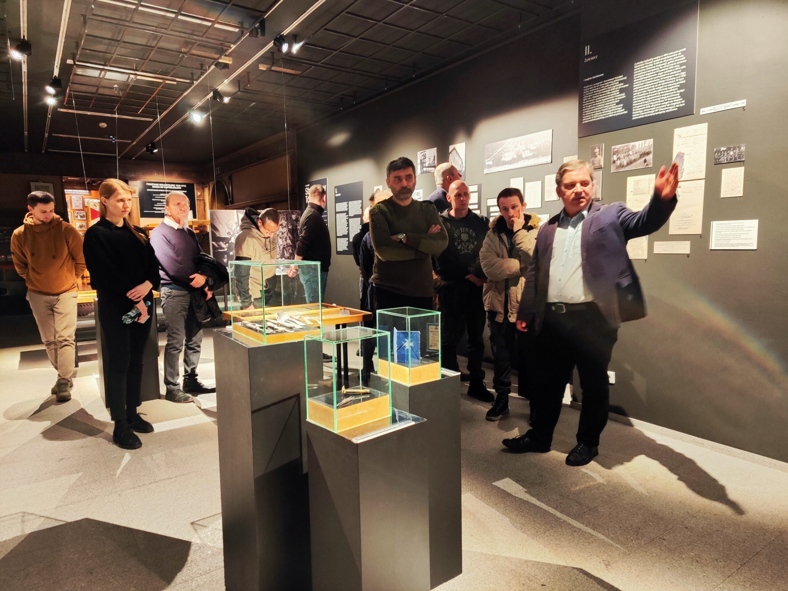 Zdjęcie przedstawia grupę osób zwiedzających wystawę czasową o Stanisławie Taczaku „Naukowiec, dowódca, społecznik” w Muzeum Powstania Wielkopolskiego 1918-1919. Na pierwszym planie przewodnik, który wskazuję ręką na konkretną fotografię z życia bohatera.