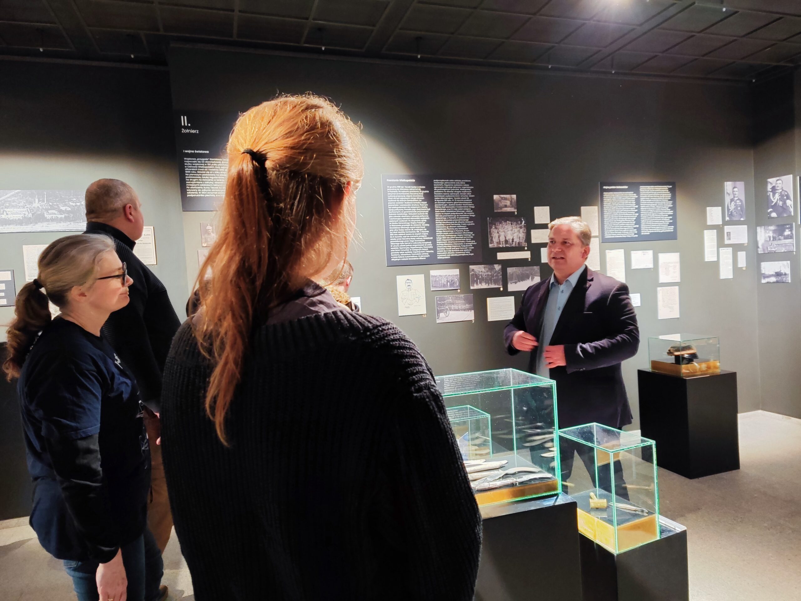 Zdjęcie przedstawia grupę osób zwiedzających wystawę czasową „Generał Stanisław Taczak - naukowiec, dowódca, społecznik” w Muzeum Powstania Wielkopolskiego 1918-1919 z przewodnikiem.