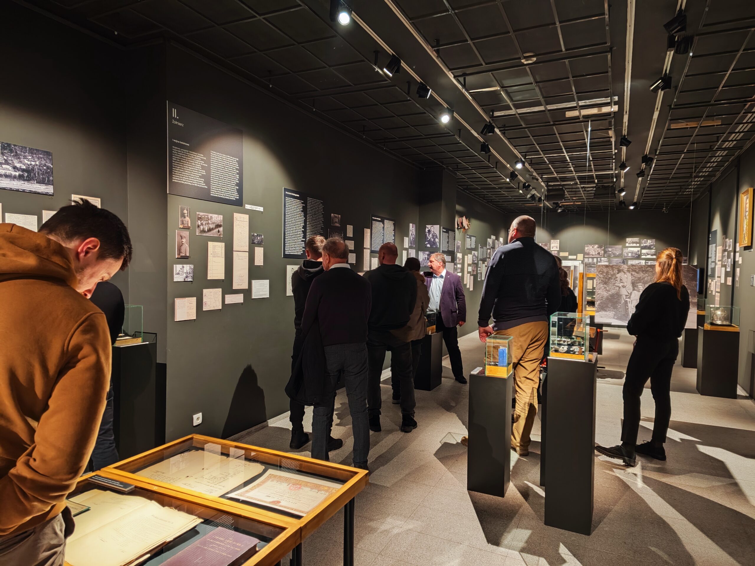 Zdjęcie przedstawia grupę osób zwiedzających wystawę czasową „Generał Stanisław Taczak - naukowiec, dowódca, społecznik” w Muzeum Powstania Wielkopolskiego 1918-1919 z przewodnikiem.