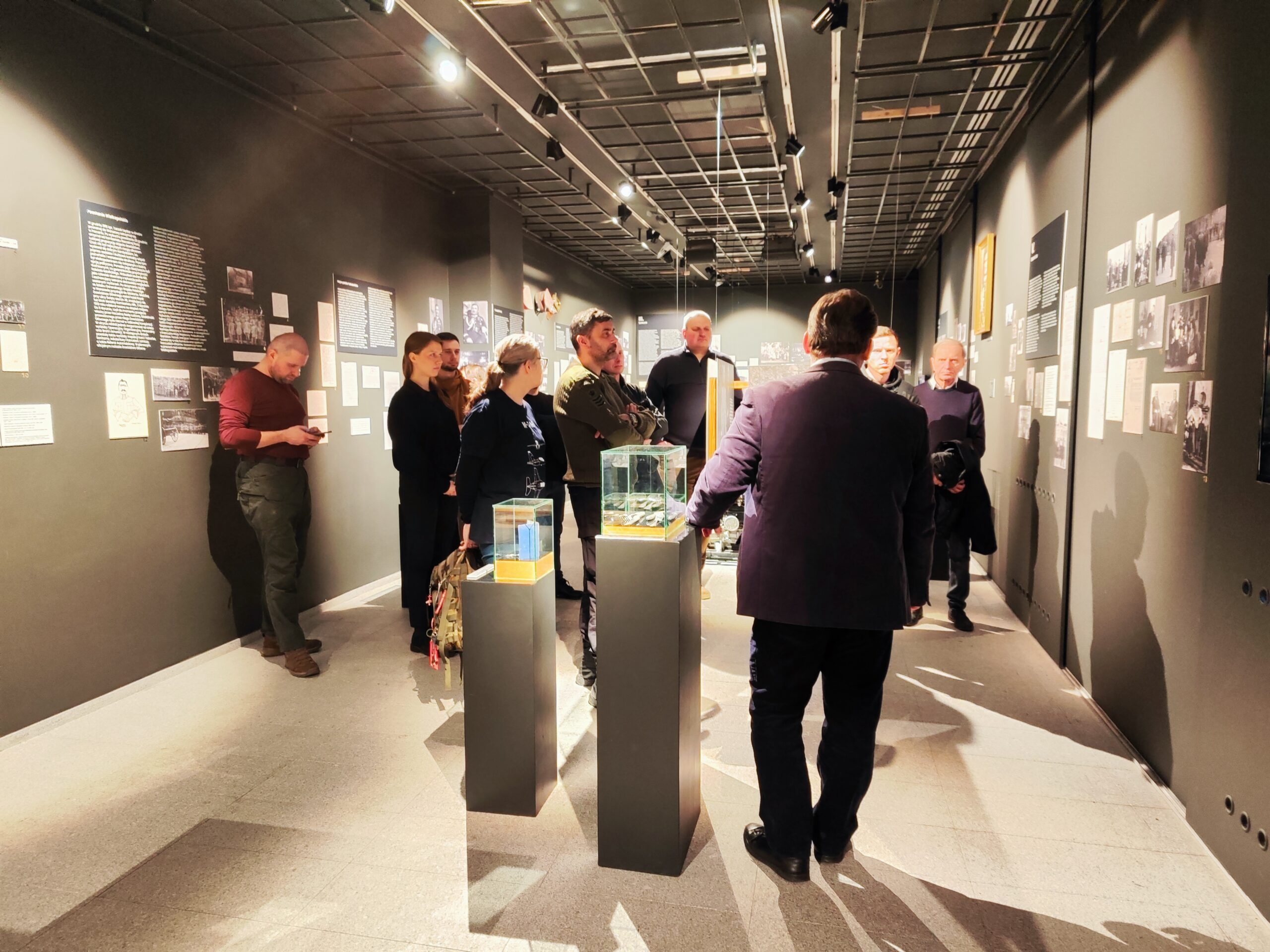 Zdjęcie przedstawia grupę osób zwiedzających wystawę czasową „Generał Stanisław Taczak - naukowiec, dowódca, społecznik” w Muzeum Powstania Wielkopolskiego 1918-1919 z przewodnikiem.